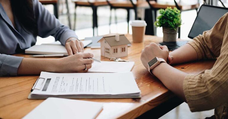 A person reviews financial documents with a pen beside a home loan application and a laptop on a kitchen table.